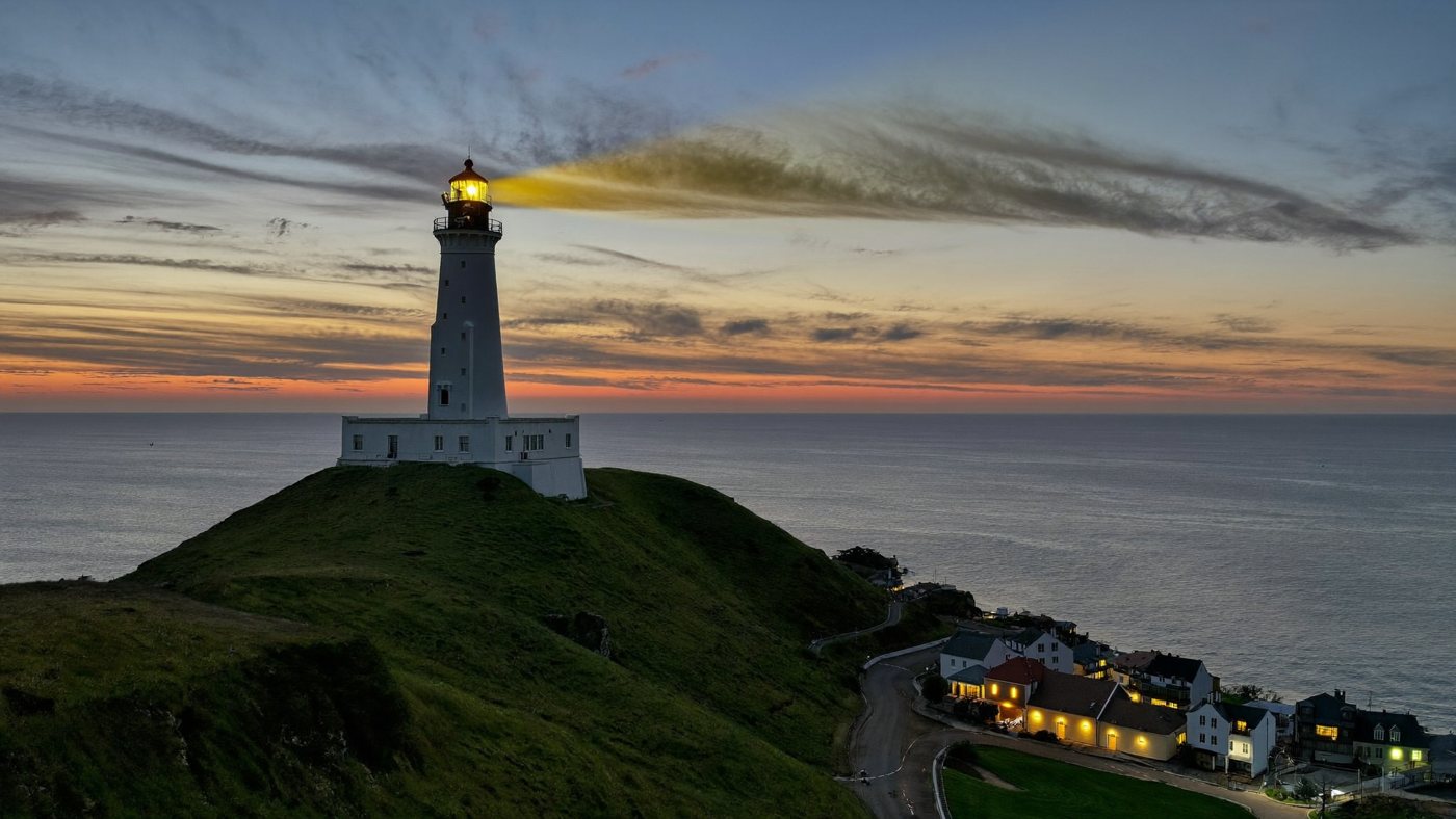 A lighthouse at dusk shining over a small coastal town